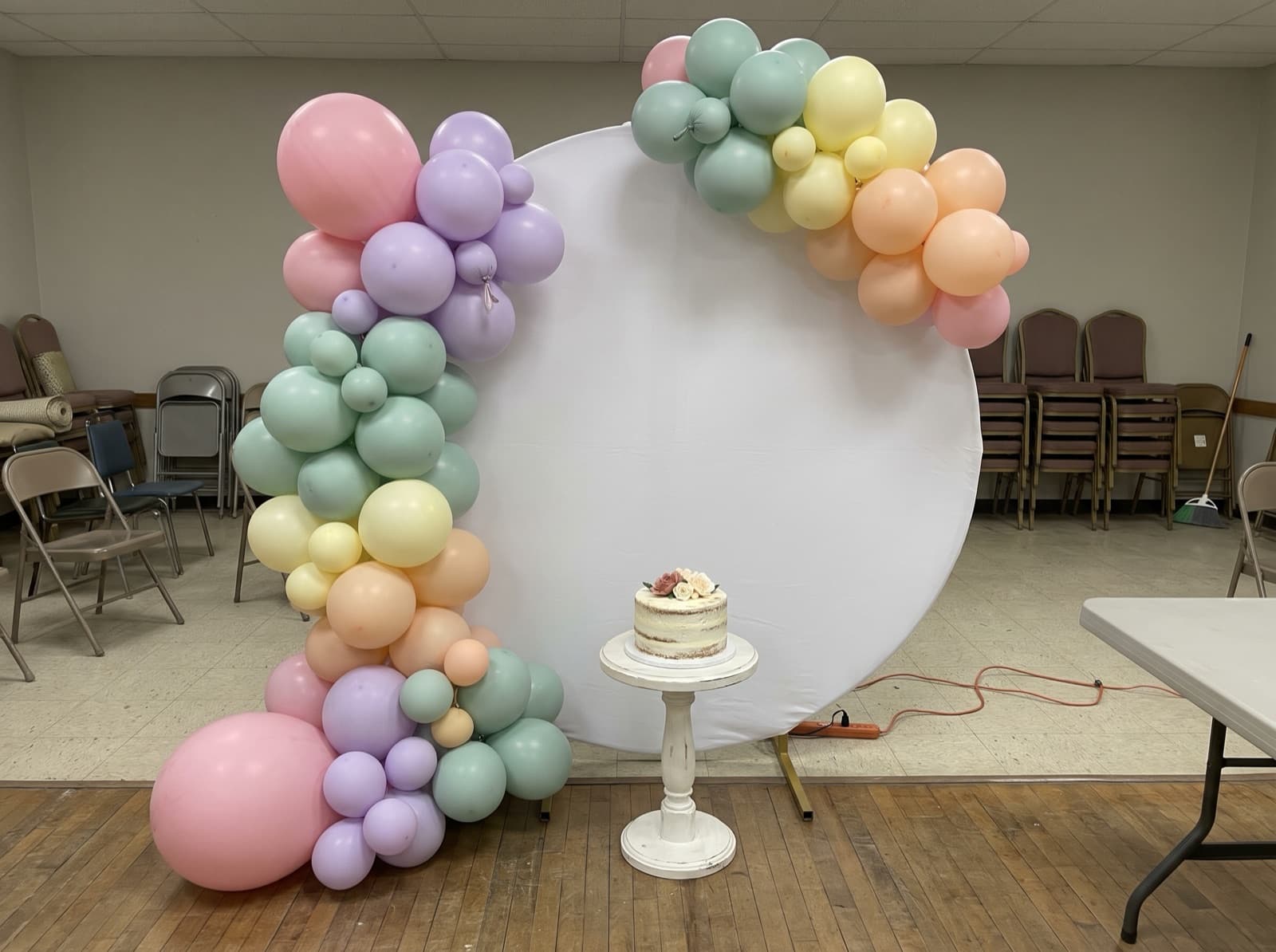 Round balloon backdrop with a pastel organic garland and a cake pedestal at a birthday party